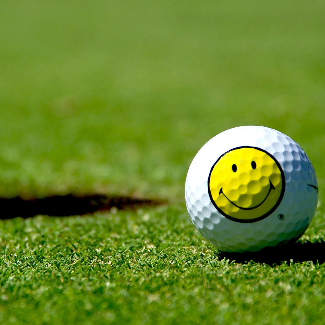 Ein Golfball mit einem gelben Smiley-Gesicht in der Nähe eines Lochs auf einem grünen Golfplatz. Die Szene vermittelt eine spielerische, fröhliche Stimmung.