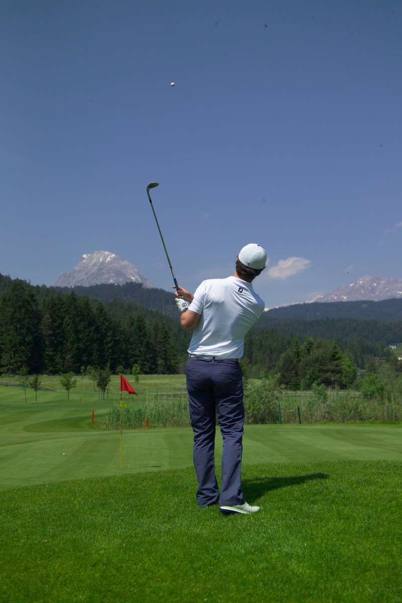 Ein Golfer mit weißer Kappe und weißem Hemd schwingt seinen Schläger auf einem üppig grünen Golfplatz, umgeben von Bäumen und fernen Bergen unter einem strahlend blauen Himmel.