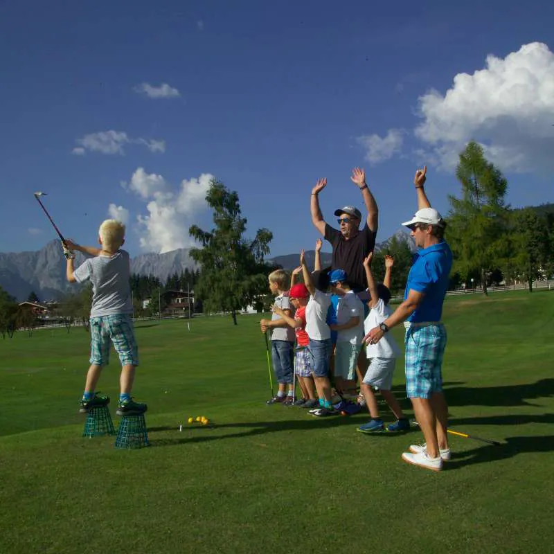 Ein Junge balanciert auf Kisten und schwingt an einem sonnigen Tag einen Golfschläger. Eine Gruppe von Kindern und zwei Erwachsene jubeln mit erhobenen Armen und vermitteln Begeisterung und Ermutigung auf dem Golfplatz.