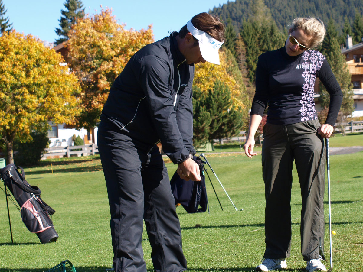 Ein Mann in einem schwarzen Trainingsanzug demonstriert einer lächelnden Frau an einem sonnigen Tag einen Golfschwung. Im Hintergrund sind herbstliche Bäume und Golftaschen zu sehen.