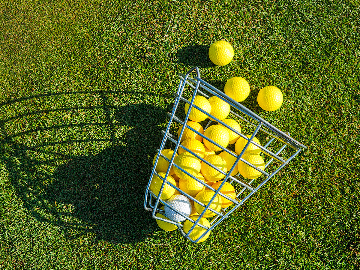 Ein Metallkorb mit gelben Golfbällen und einem weißen Ball liegt umgekippt auf dem Rasen und wirft einen Schatten. Die Szene vermittelt eine ungezwungene, sonnige Stimmung.