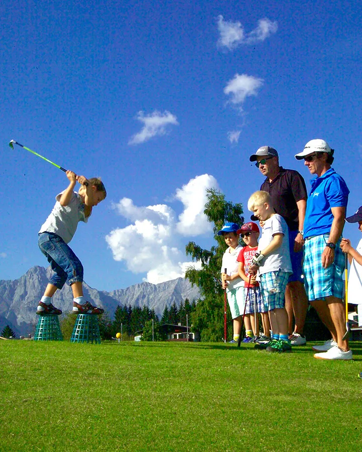 Ein kleines Kind schwingt voller Energie einen Golfschläger auf dem Grün, während andere, darunter auch Erwachsene, interessiert zuschauen. Die Sonne scheint, im Hintergrund sind Berge zu sehen.