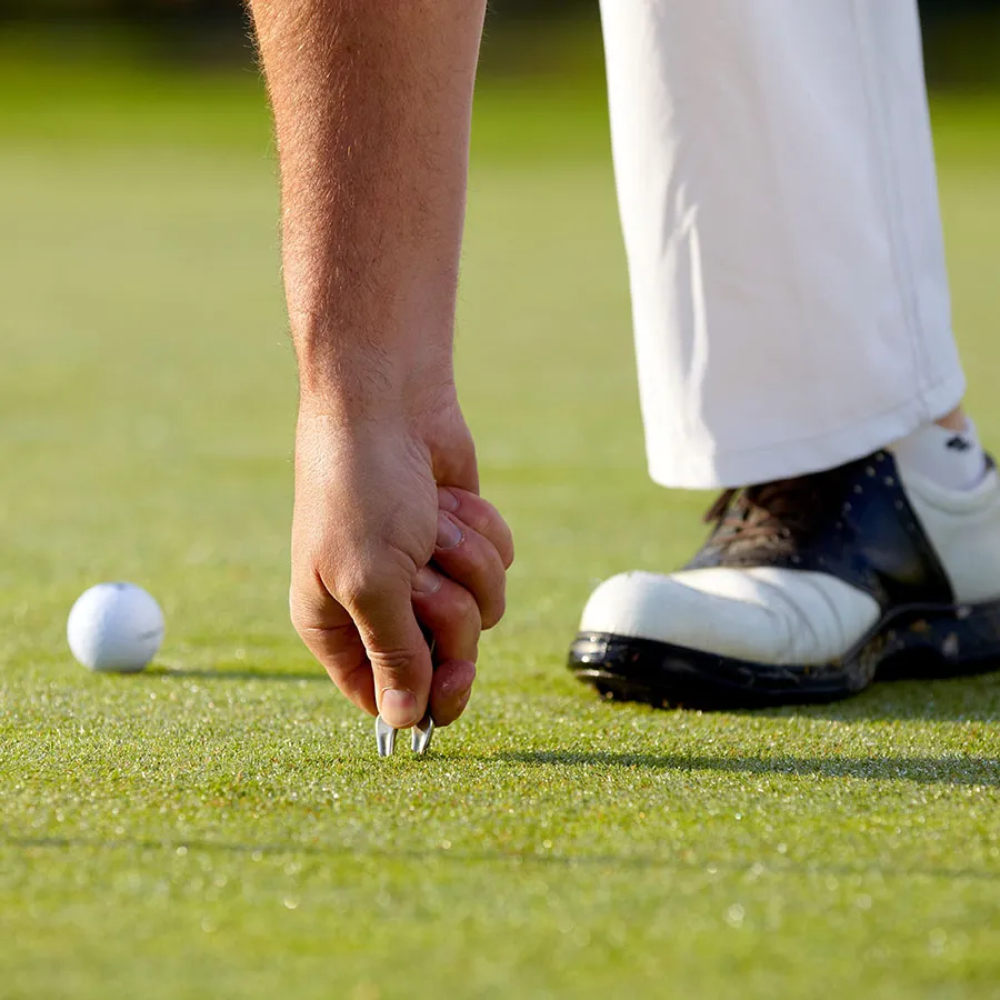 Die Hand eines Golfers repariert eine Grasnarbe auf einer üppig grünen Putting-Fläche. In der Nähe liegt ein Golfball, und der Golfer trägt weiße Schuhe und Hosen. Die Szene ist ruhig und konzentriert.